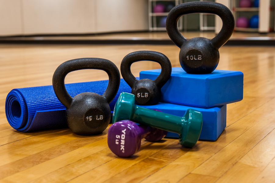 Various workout equipment sit on a wood floor: kettle bells, yoga mat, plastic weights and bricks