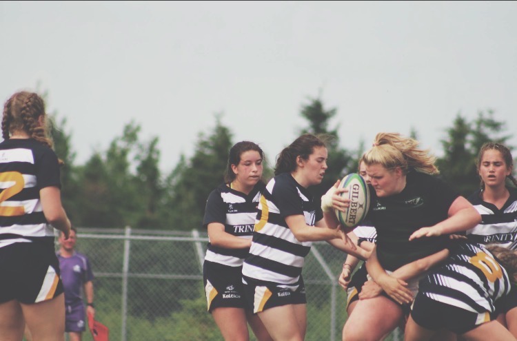 A woman in a black uniform holding a rugby ball charges through a group of women wearing striped uniforms