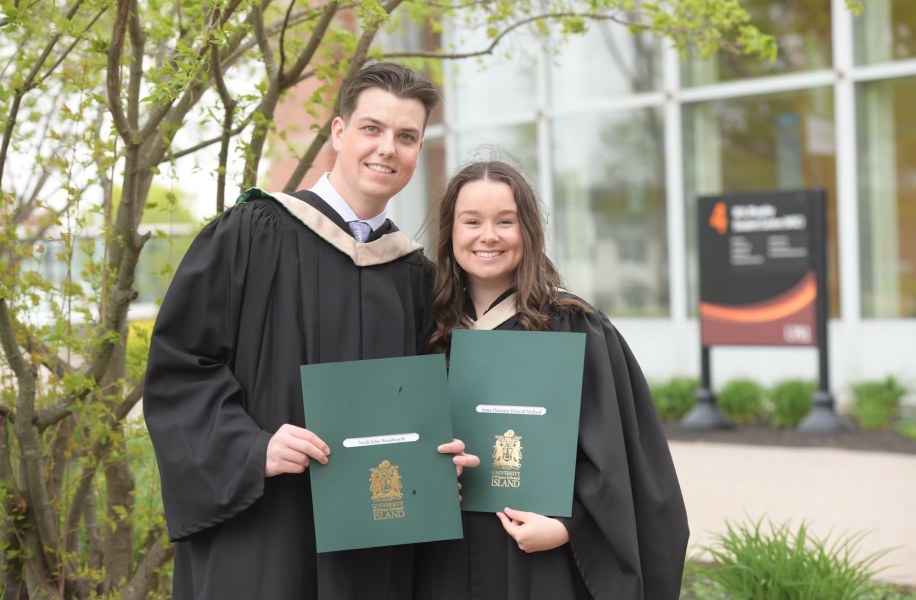 A smiling male and female wearing academic robes clutch embossed folders with the crest of UPEI on the cover