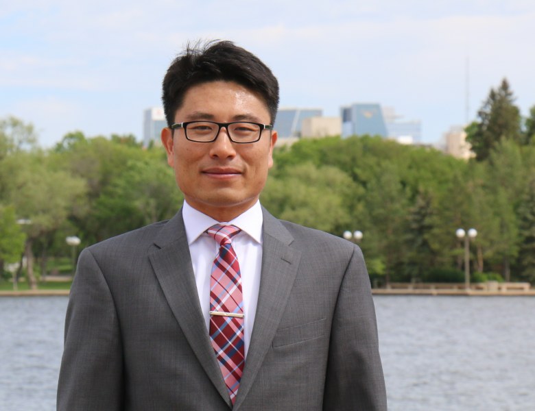 A smartly dressed man in a grey suit stands in front of a river with distant buildings in the background