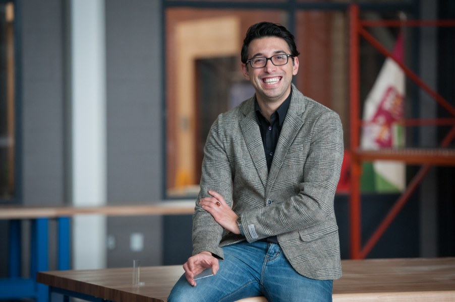 A smiling spectacled man in a casual blazer and jeans