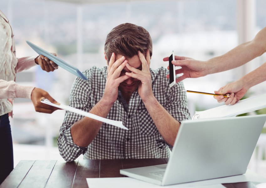 Man sitting at desk holding his head in his hands surrounded by stressors