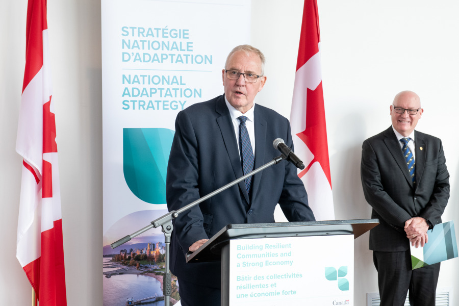 man speaking at podium flanked by two Canadian flags with man looking on