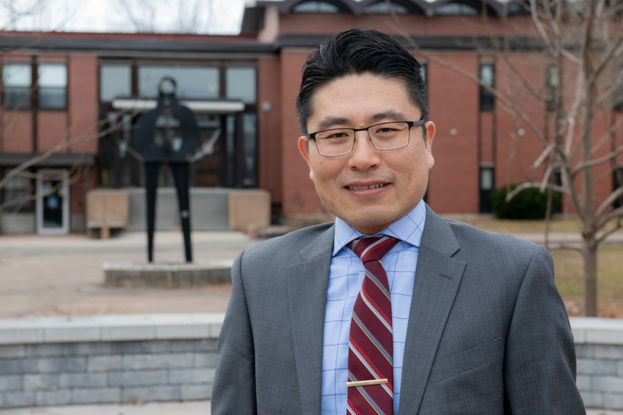 photo of man wearing glasses standing in front of a metal sculpture and red brick building