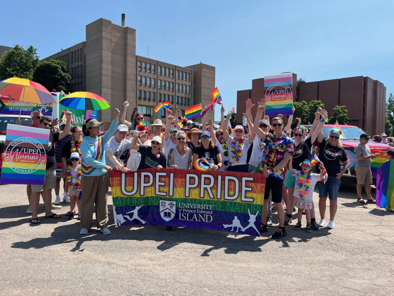 Members of the UPEI community at the 2024 PEI Pride Parade Photo of large group of people holding a banner and wearing clothing in bright rainbow colours