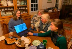 Two female students sit around a farm table speaking with an older couple