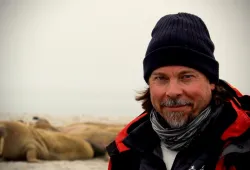 A medium close-up shot of a bearded man in winter hat and jacket in front of a chilly landscape