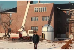 An aged photograph of a man standing next to a silver dome on the ground next to a brick building