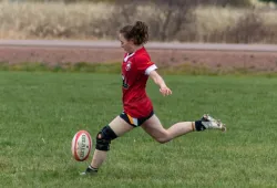 A female athlete extends her leg to kick a rugby ball on a grassy pitch