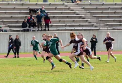 A female rugby player in a green UPEI Panthers jersey runs up a field with a look of determination in her eyes. She is trailed by competing players from Saint Mary's University. 