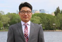 A professional-looking man n a grey suit stands in front of a river with trees and buildings on the opposite shore