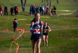 A woman running in a long distance race, looking tired but strong. The background shows many runners behind her on a grassy course with a dirt path. 