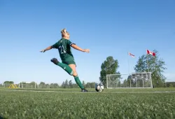 A female soccer player in a green Panthers soccer uniform winds up with her right foot to take a shot on net