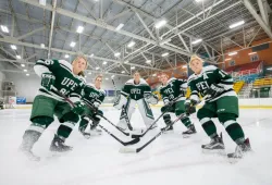 Five women in skates and green Panthers hockey gear on the ice. A goalie stands motionless in the middle. Four other players have obviously just staked into frame