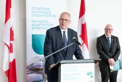 man speaking at podium flanked by two Canadian flags with man looking on