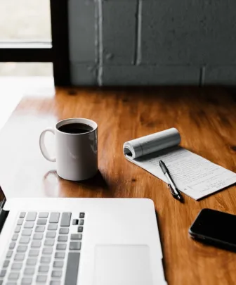 wooden table with a laptop computer, cup of coffee, cellular phone, and notepad